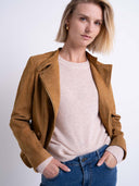 A woman wearing a tan suede moto jacket over a light pink knit sweater and blue jeans, photographed against a clean white studio background.