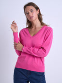 A woman modeling a bright pink long-sleeve V-neck knit sweater, paired with dark blue jeans, against a light gray studio background.