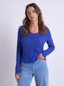 A woman modeling a bright blue cable-knit long-sleeve top with a scalloped hem, paired with light-wash blue jeans, against a plain light gray studio background.