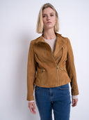A woman modeling a tan suede moto jacket with gold-tone hardware, paired with a cream knit top and blue jeans, against a plain light gray studio background.
