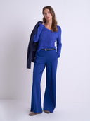 A woman modeling a royal blue knit top and matching wide-leg trousers with a black belt, holding a dark navy jacket over her shoulder, against a light gray studio background.