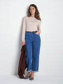 A woman stands against a plain white studio wall wearing a beige long-sleeve top, blue wide-leg jeans with rolled cuffs, and brown shoes, while holding a brown coat.