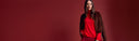 A woman modeling a dark brown faux-fur jacket over a bright red mock-neck sweater and matching red skirt, standing against a solid dark red studio background.