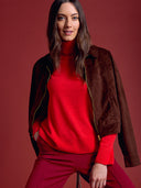 A woman wearing a brown suede jacket over a bright red turtleneck sweater and matching red trousers, posed against a solid dark red studio background.