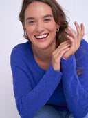A smiling woman with brown hair wearing a bright blue textured knit sweater and light blue jeans, posing against a light gray studio background.