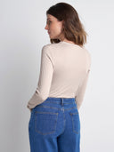 A woman shown from the back wearing a beige long-sleeve top and blue jeans against a plain light gray studio background.