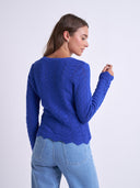 A woman shown from the back wearing a royal blue cable-knit sweater with a scalloped hem and light blue jeans, photographed against a plain light gray studio background.