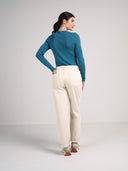 A woman is shown from the back wearing a teal and navy blue horizontally striped long-sleeve top, cream-colored straight-leg trousers, and beige sneakers, set against a plain light gray studio background.