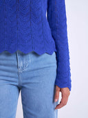 A close-up of a person wearing a bright royal blue textured knit sweater with a scalloped hem and light blue jeans against a plain light background.