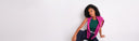 A woman with curly black hair sits against a plain white wall wearing a bright pink open-front blazer over a dark green top and dark blue pants.
