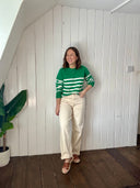 Woman wearing a green and white striped sweater and beige pants standing against a white paneled wall.